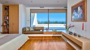 A modern bedroom with a large window, a bookshelf, a sofa, and a bedside table with decorative items. - Villa Anastasia - Private Pool Panoramic Views (Platanias)