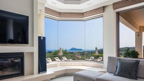A modern living room with a flat-screen TV, a fireplace, and a sofa with a view of the landscape. - Villa Anastasia - Private Pool Panoramic Views (Platanias)