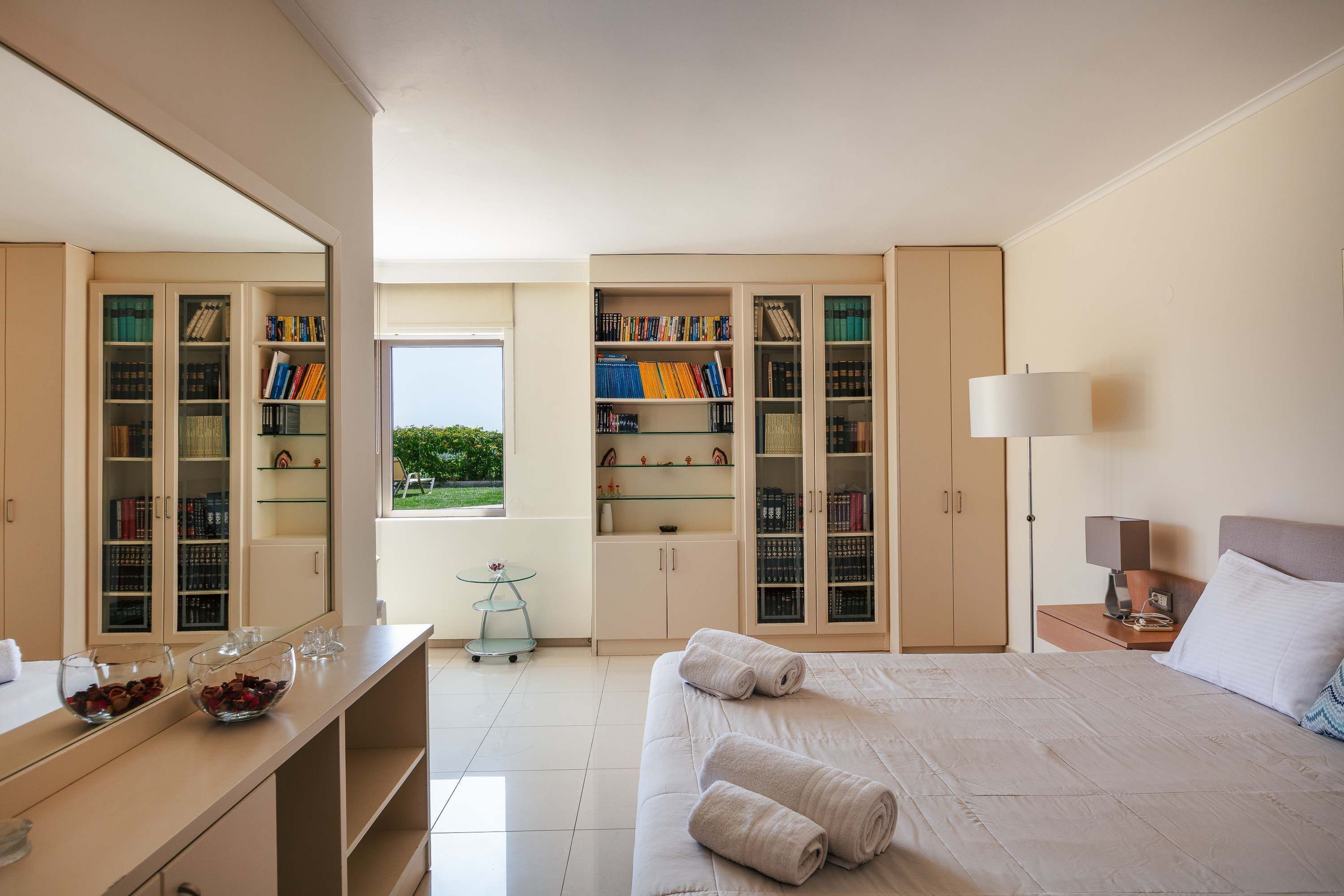 A hotel room with a large bed, a desk with a bowl of chocolates, a bookshelf, and a window with a view of greenery.