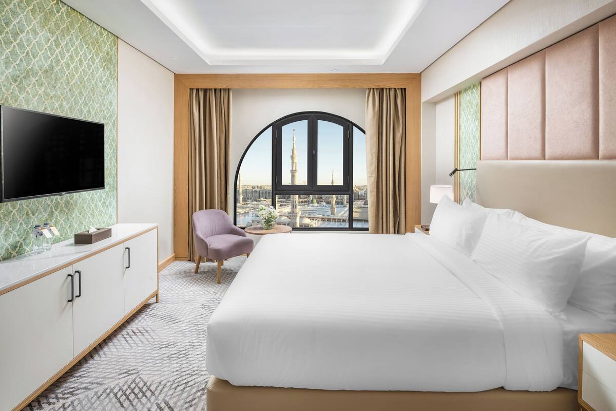 Hotelkamer met een groot bed, een televisie links, uitzicht op de stad door een groot raam en een klein tafeltje met een stoel.