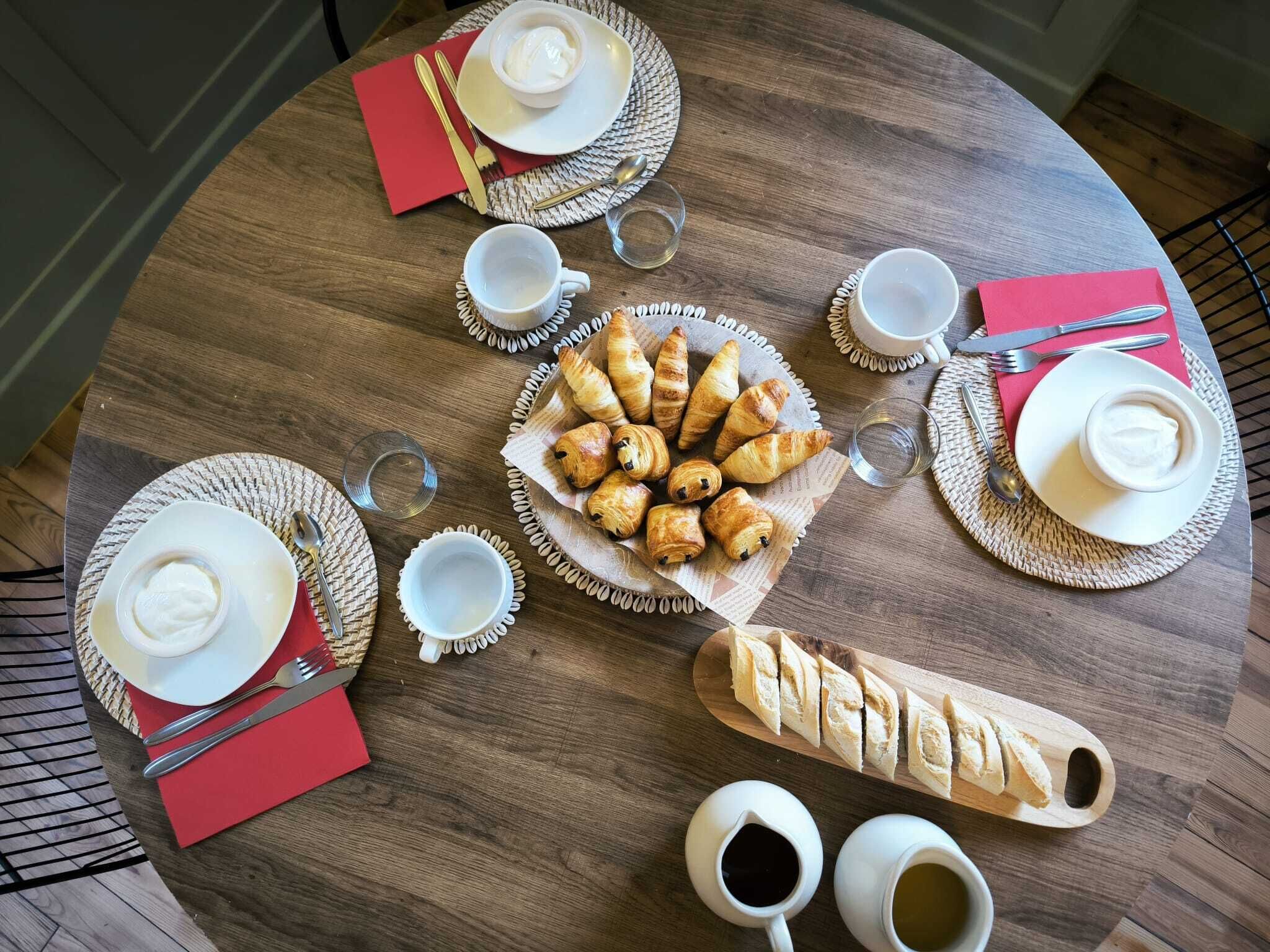 A round wooden table set with plates, croissants, bread, and cups, suggesting a breakfast setting.
