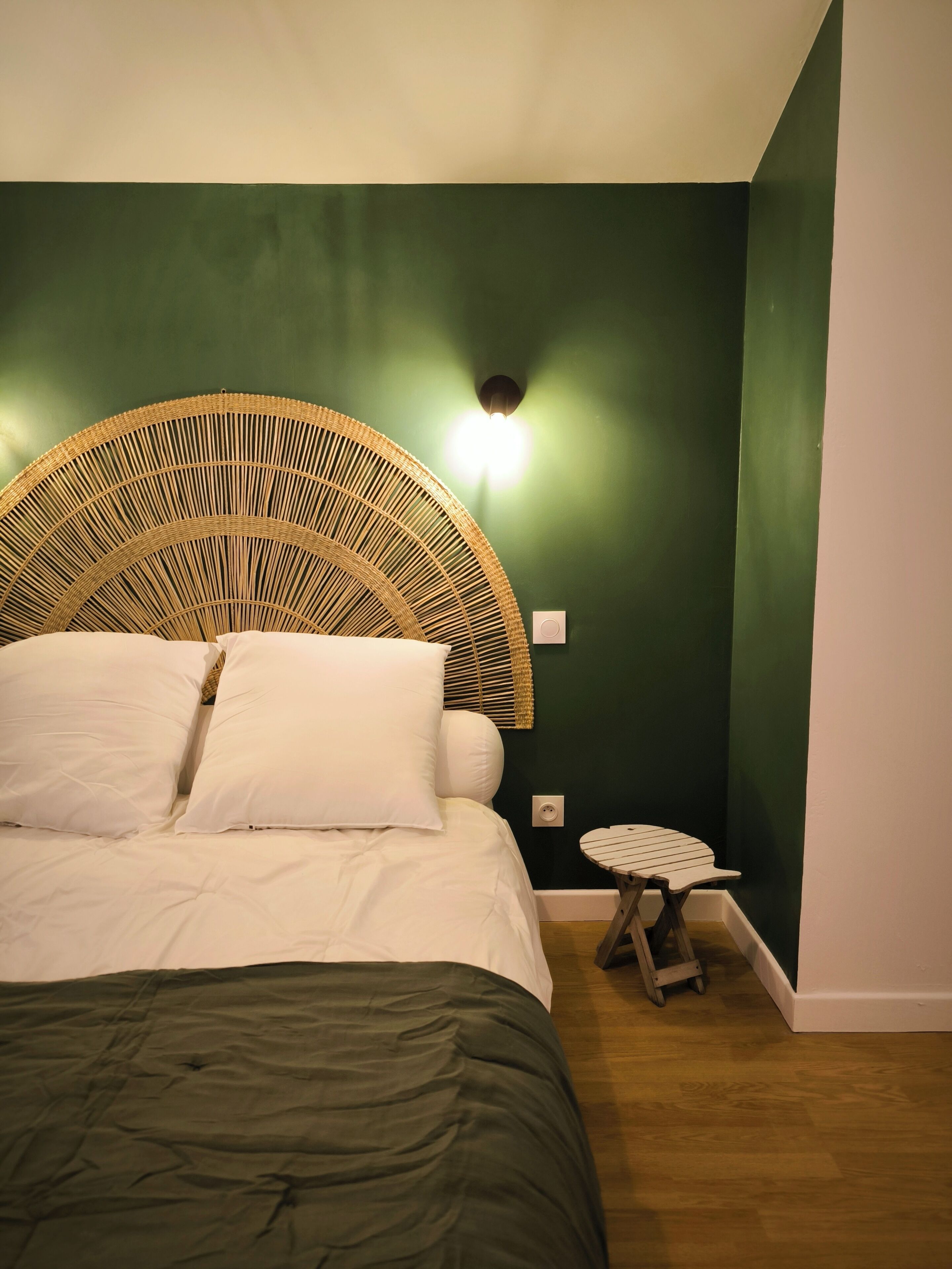 A bedroom with a large bed, a wicker headboard, a small table, and a wall-mounted light.