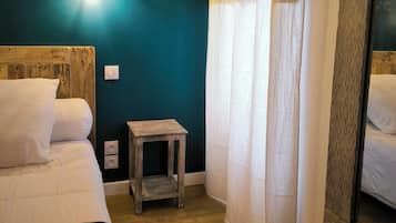 Un dormitorio con cabecera de madera, un taburete pequeño de madera, una cama con ropa blanca y una pared color aguamarina, además de un espejo grande.