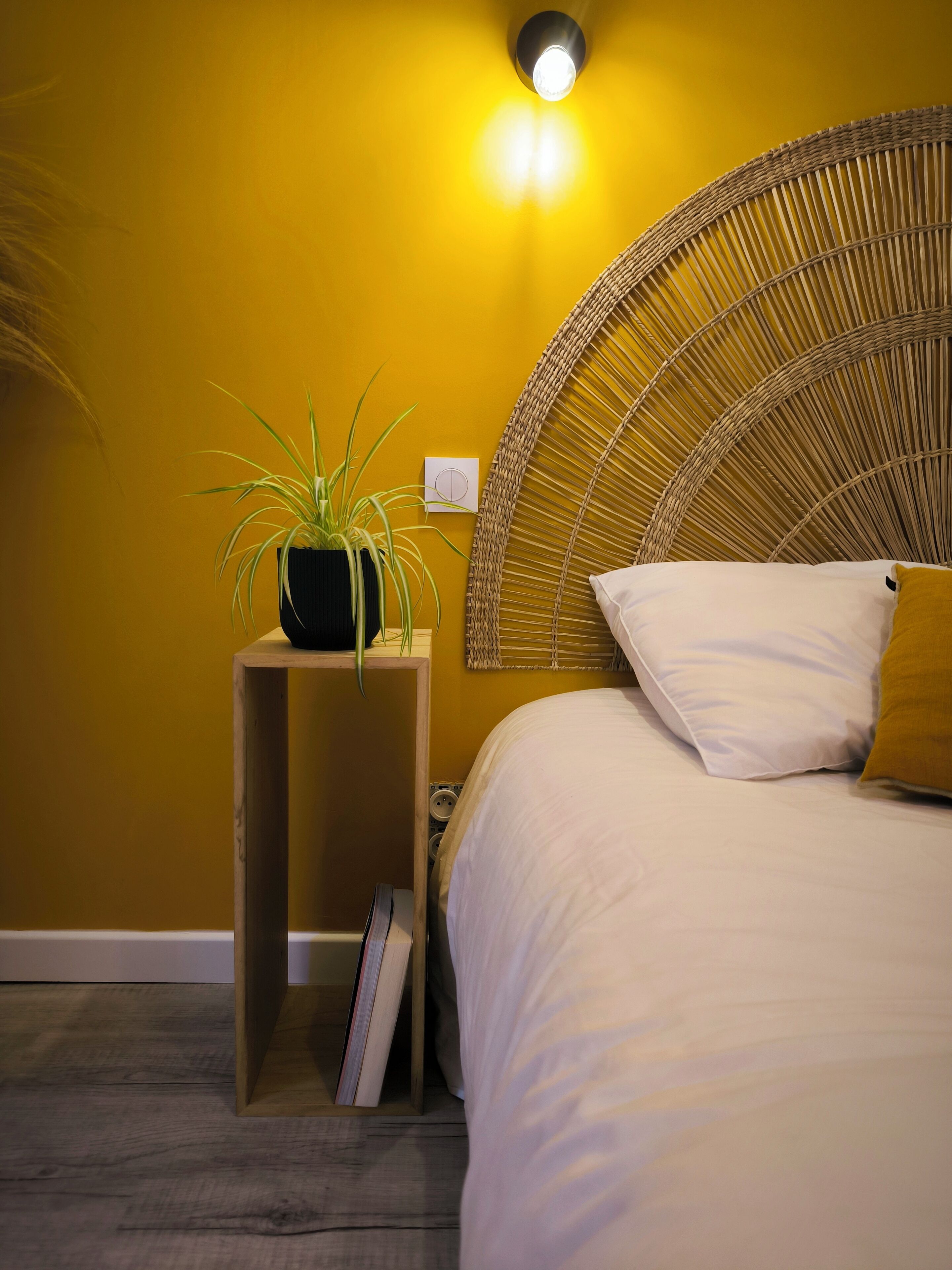Ein Schlafzimmer mit einem Bett, einem Nachttisch mit Pflanze, einer Wandleuchte und einem gewobenen Kopfteil.