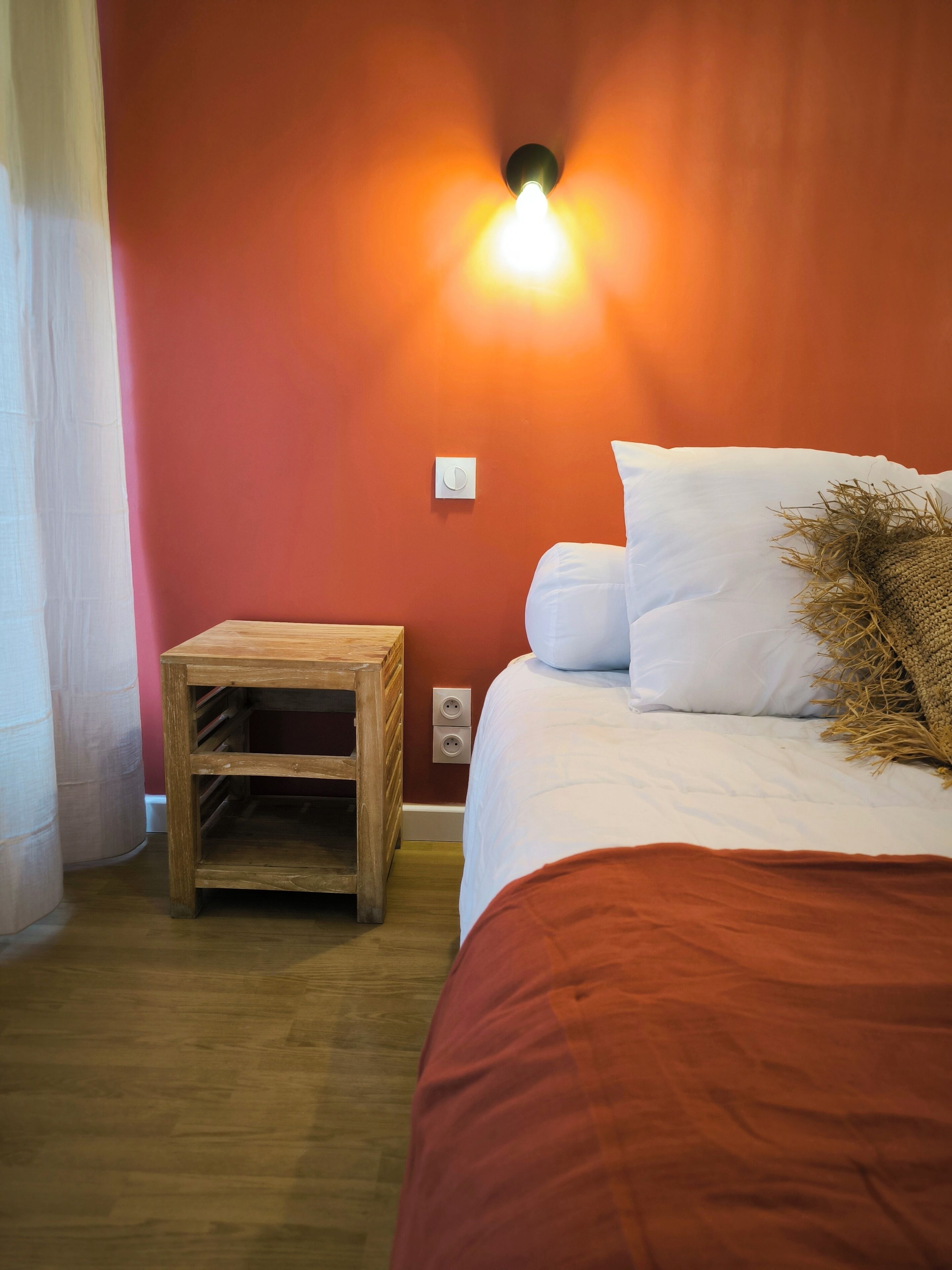 Un dormitorio con una mesita de noche de madera, una cama con ropa de cama blanca, una pared roja y un aplique de pared.