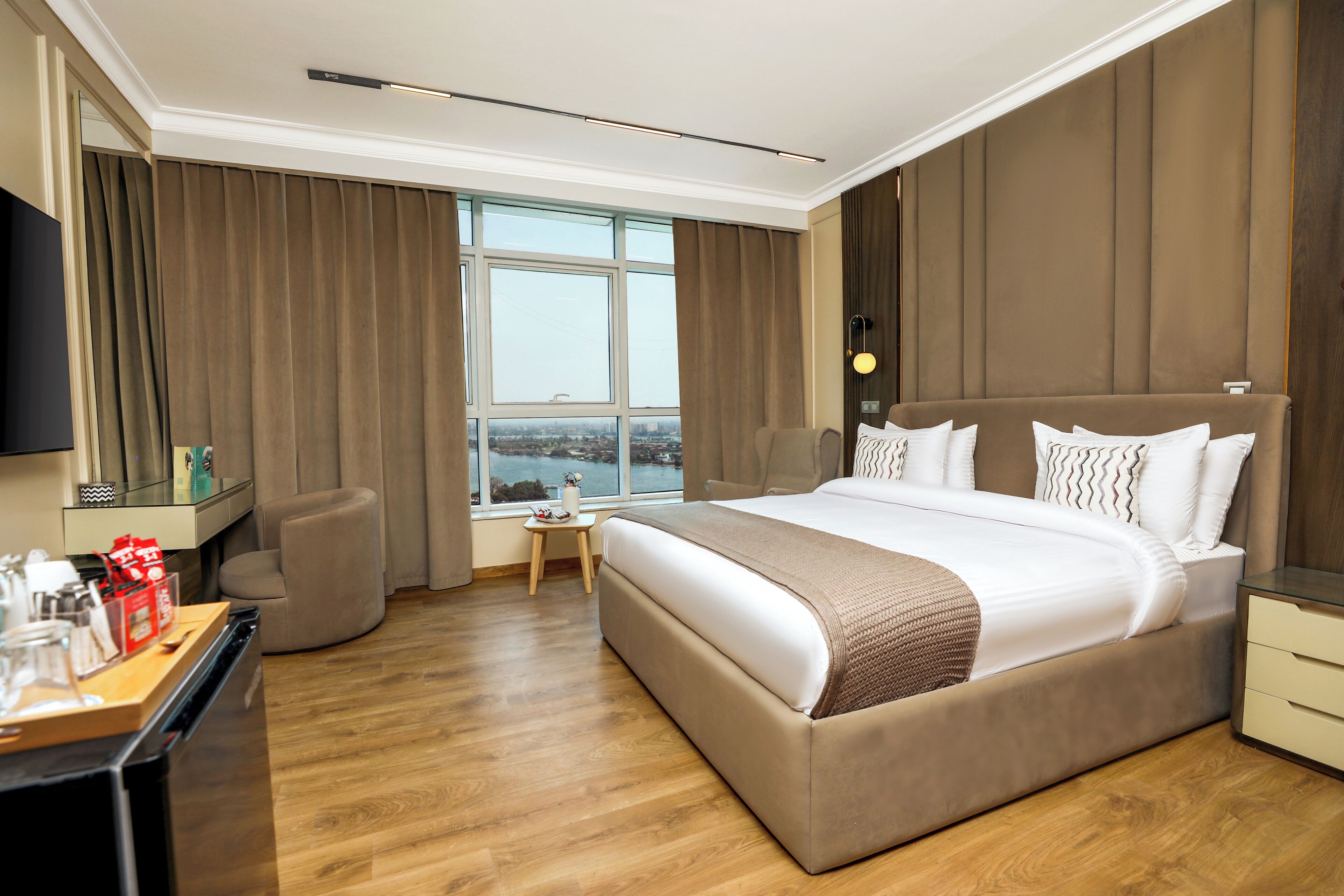 Una habitación de hotel moderna con una cama grande, un escritorio con una silla, una mesita con una lámpara y un ventanal con vistas a la ciudad.