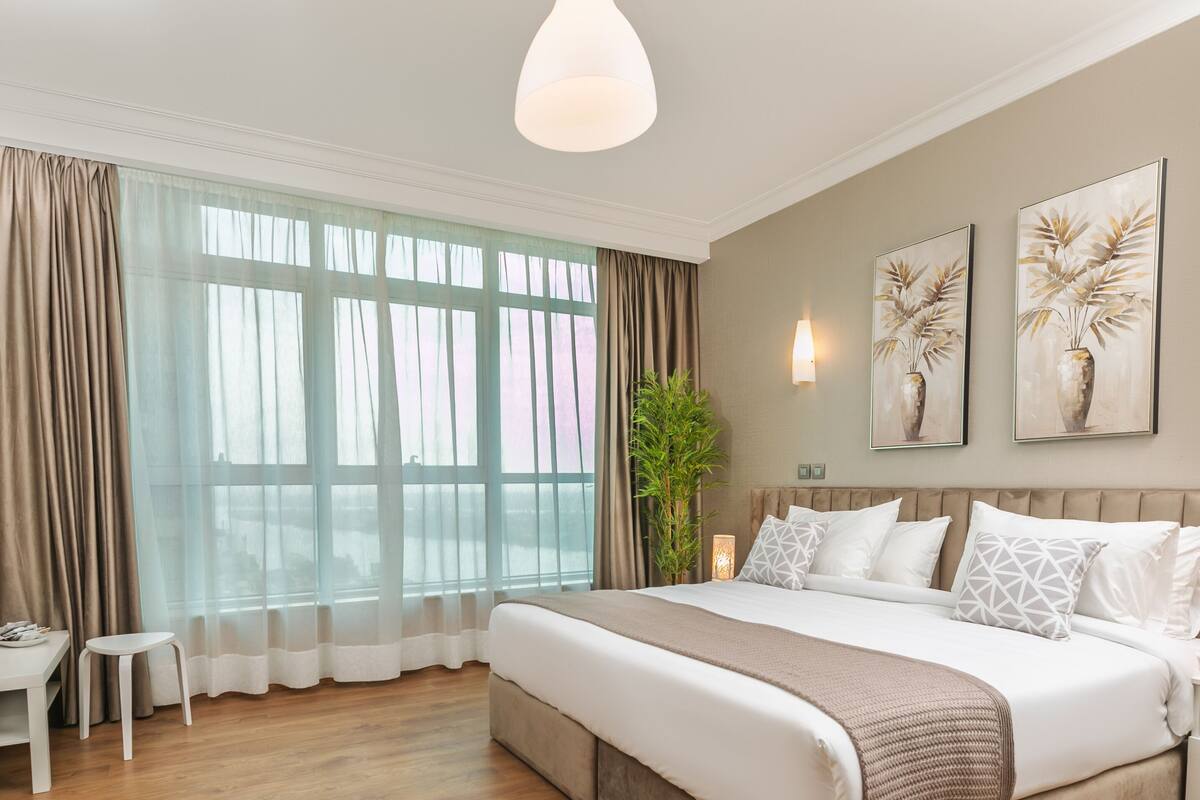 A modern hotel room with a large bed, a TV, a small table, and a large window with curtains.