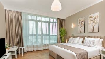 Una habitación de hotel moderna con una cama grande, televisor, una mesita pequeña y un ventanal con cortinas.