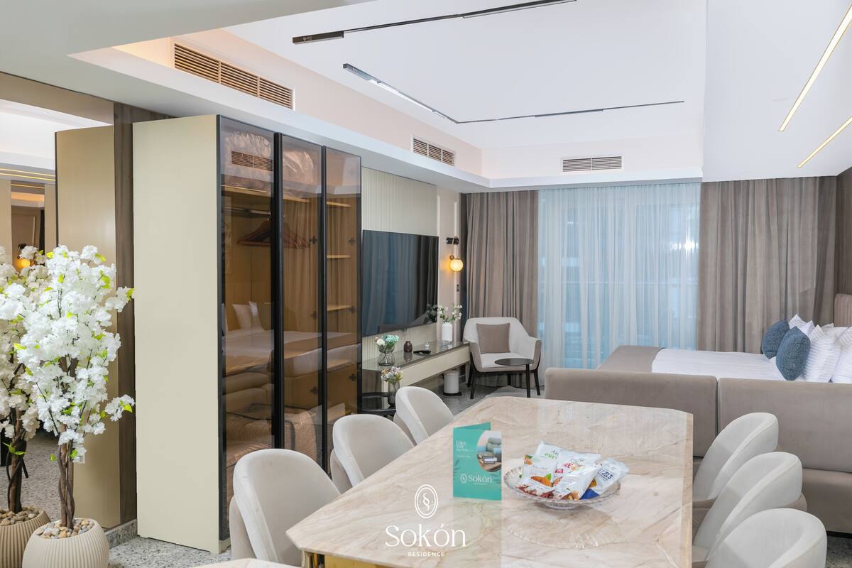 A modern hotel room with a dining area, a sofa, a bed, and a large window with curtains.