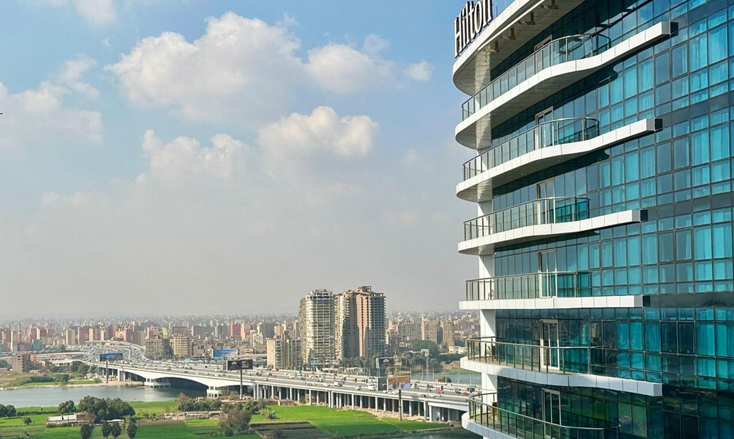 Un hotel moderno con fachada de vidrio, varios balcones y vistas a un paisaje urbano que incluye un puente y un río.