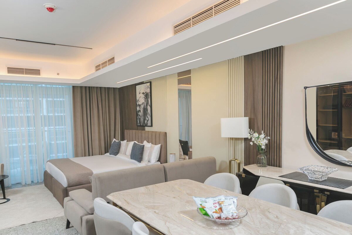 Ein modernes Hotelzimmer mit einem großen Bett, einer Sitzecke mit einer Couch und Sesseln, einem Esstisch mit einer Obstschale und einem großen Spiegel.