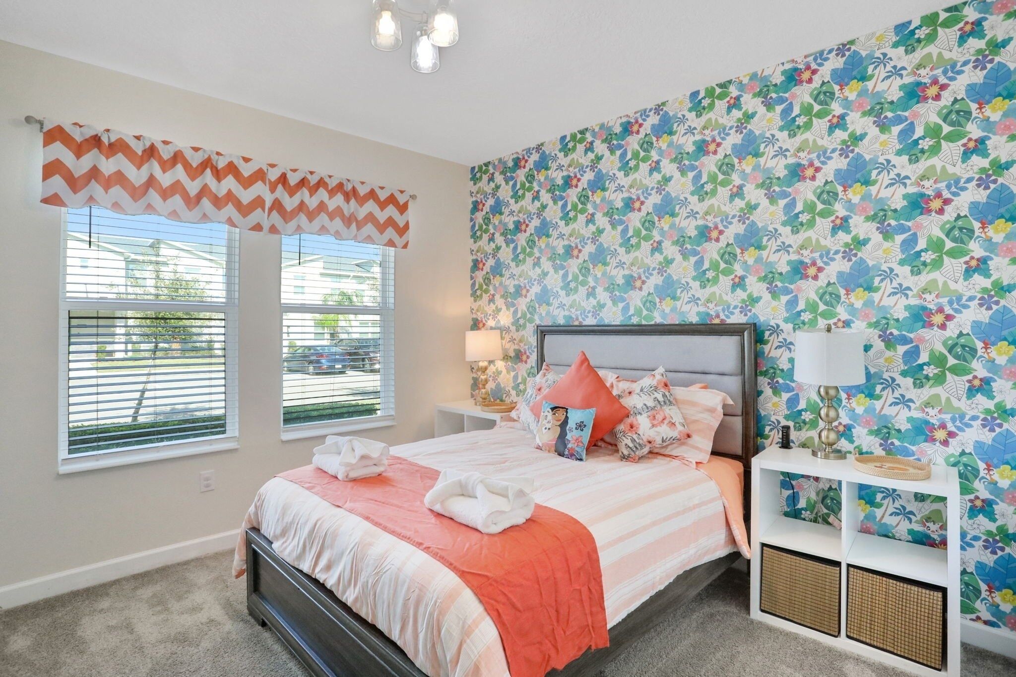 Um quarto com cama, duas janelas com persianas, papel de parede floral, mesa de cabeceira com abajur e um adorno pendurado na parede.