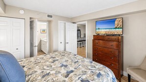 A bedroom with a bed, dresser, and a TV displaying a beach scene. - Beachfront Escape | Resort Events & Pool (St. Pete Beach)