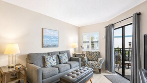 A living room with a grey sofa, patterned armchair, and a black ottoman. There are two lamps, a framed artwork on the wall, and a balcony with curtains. - Beachfront Escape | Resort Events & Pool (St. Pete Beach)