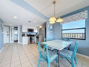 A modern kitchen with white cabinets, stainless steel appliances, and a dining area with a white table and blue chairs. - 2BR Gulf-Front | Private Beach Access (St. Pete Beach)