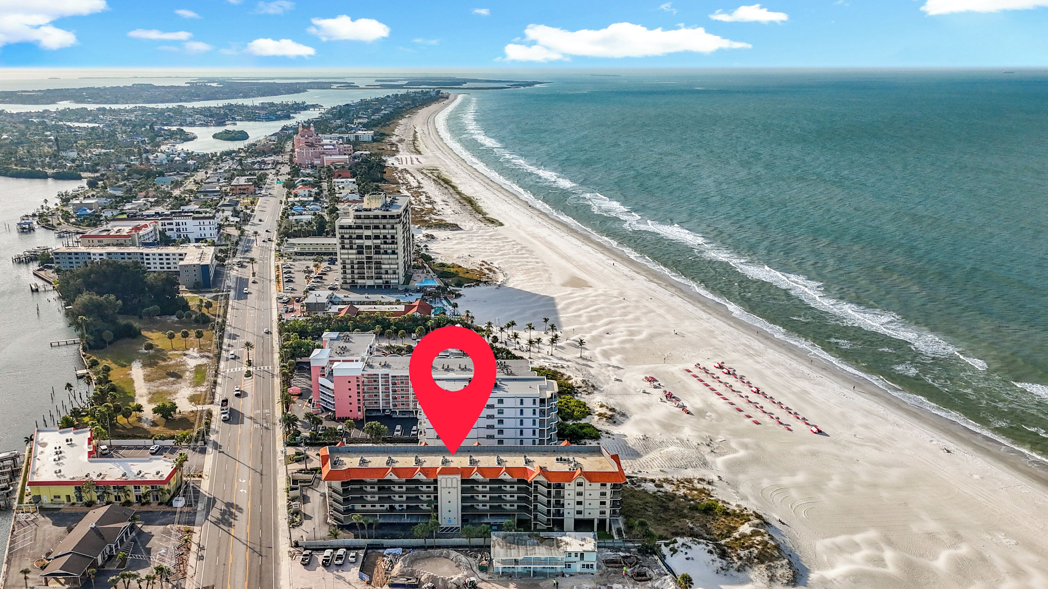 An aerial view of a coastal city with a beachfront hotel, a road parallel to the beach, and a large body of water.