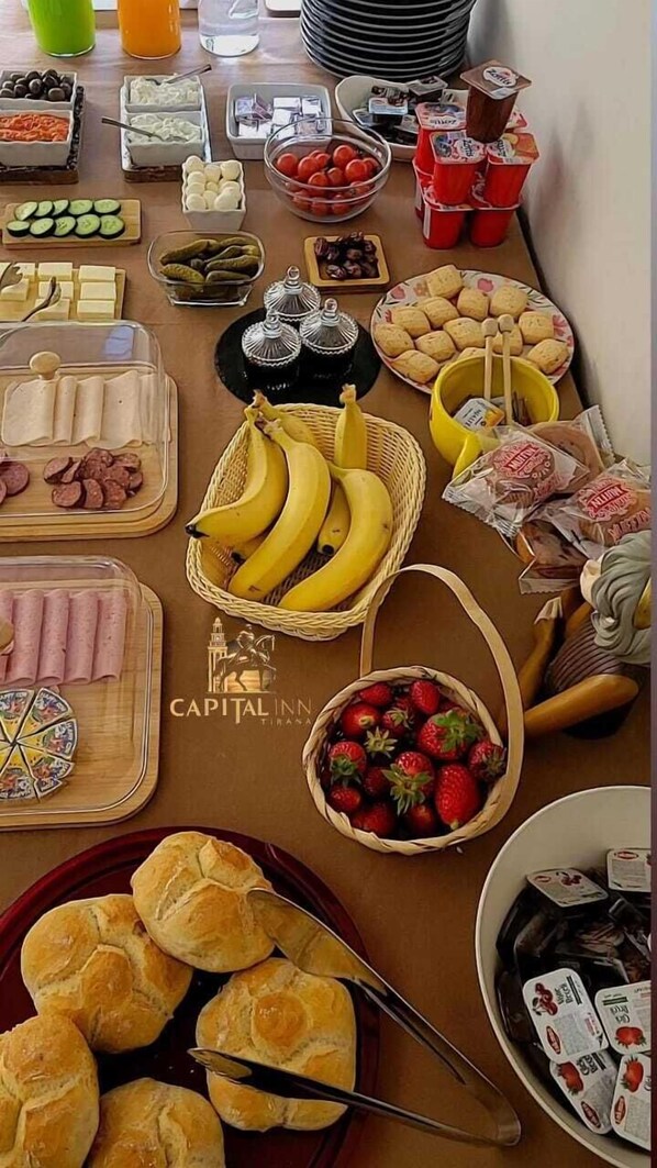 A buffet table with various food items including sliced cucumbers, tomatoes, bananas, strawberries, and bread rolls.