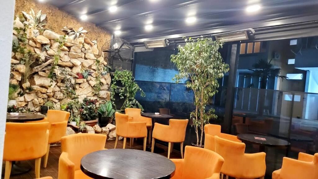An indoor seating area with round tables and orange chairs, surrounded by a rock wall and potted plants.