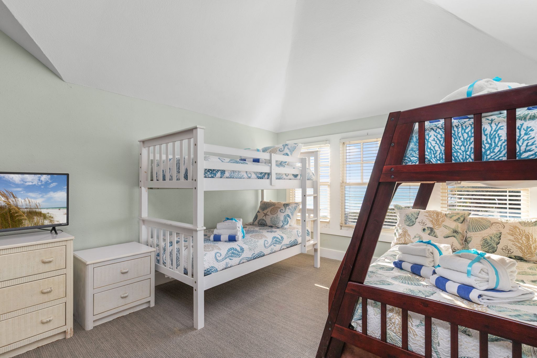 A bunk bed room with a television on a dresser, a window with blinds, and a view of the beach.