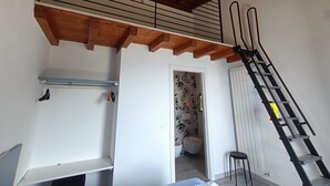 A room with a wooden ceiling, a black metal ladder, a white door, and a bathroom visible through an open doorway. - "Iseo" room (Marone)