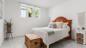Un dormitorio con una cama de madera, un pequeño cofre de madera, una planta en maceta, una mesita de noche blanca y un colgador de macramé en la pared.