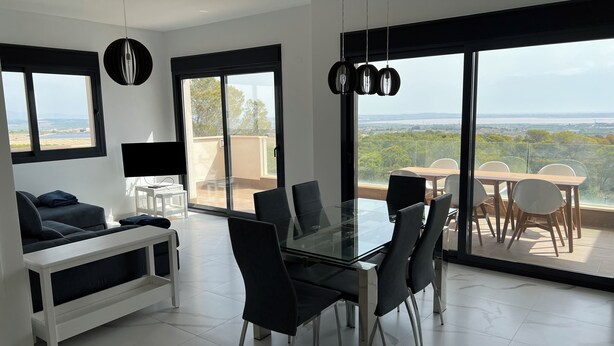 A modern dining area with a glass table and chairs, a sofa, and a TV. Large windows offer a view of the outdoors.