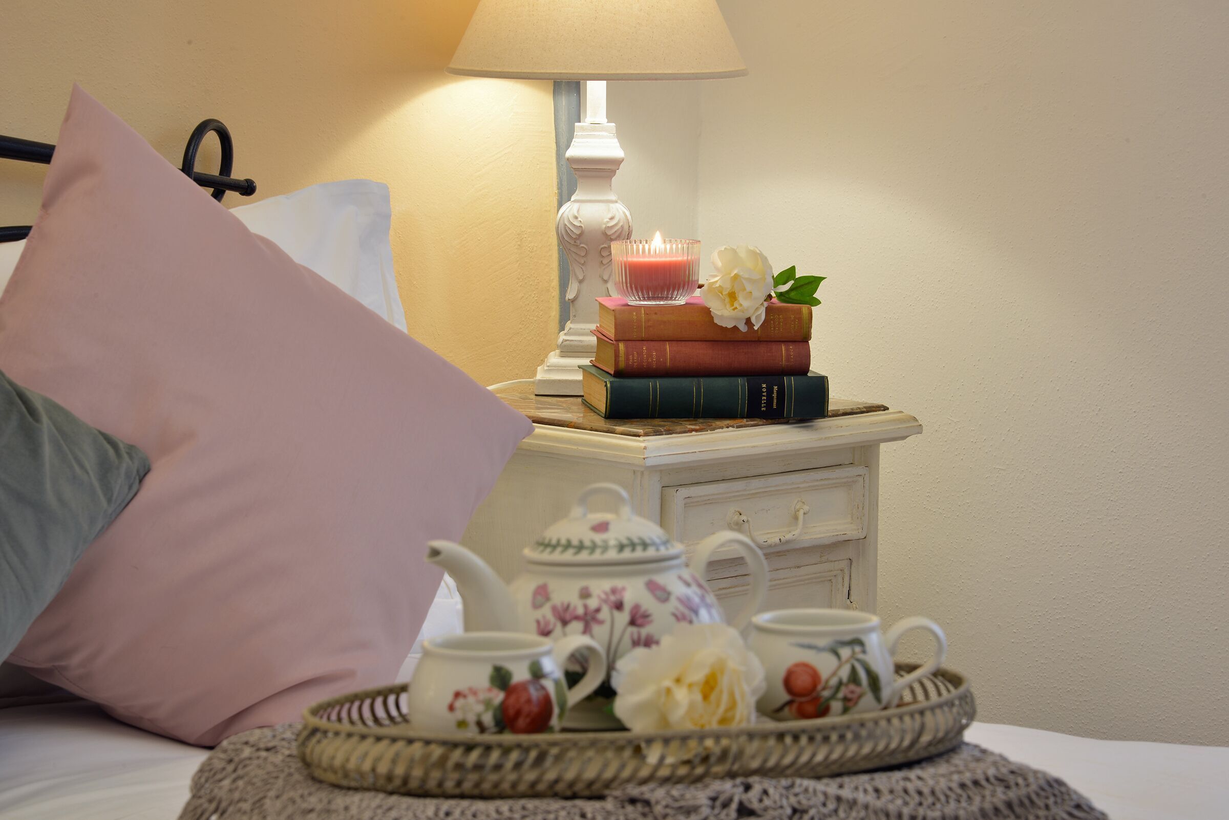 Ein ordentlich eingerichtetes Schlafzimmer mit einem Nachttisch, der eine Lampe, Bücher, eine Teekanne und ein Tablett mit Tassen und Blumen beherbergt.