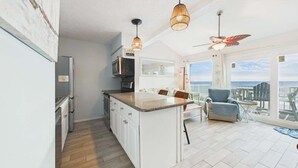 Una cocina moderna con armarios blancos, encimera de granito y una isla grande. Hay una zona de comedor con una silla azul y una mesa, además de un balcón con vistas al océano.