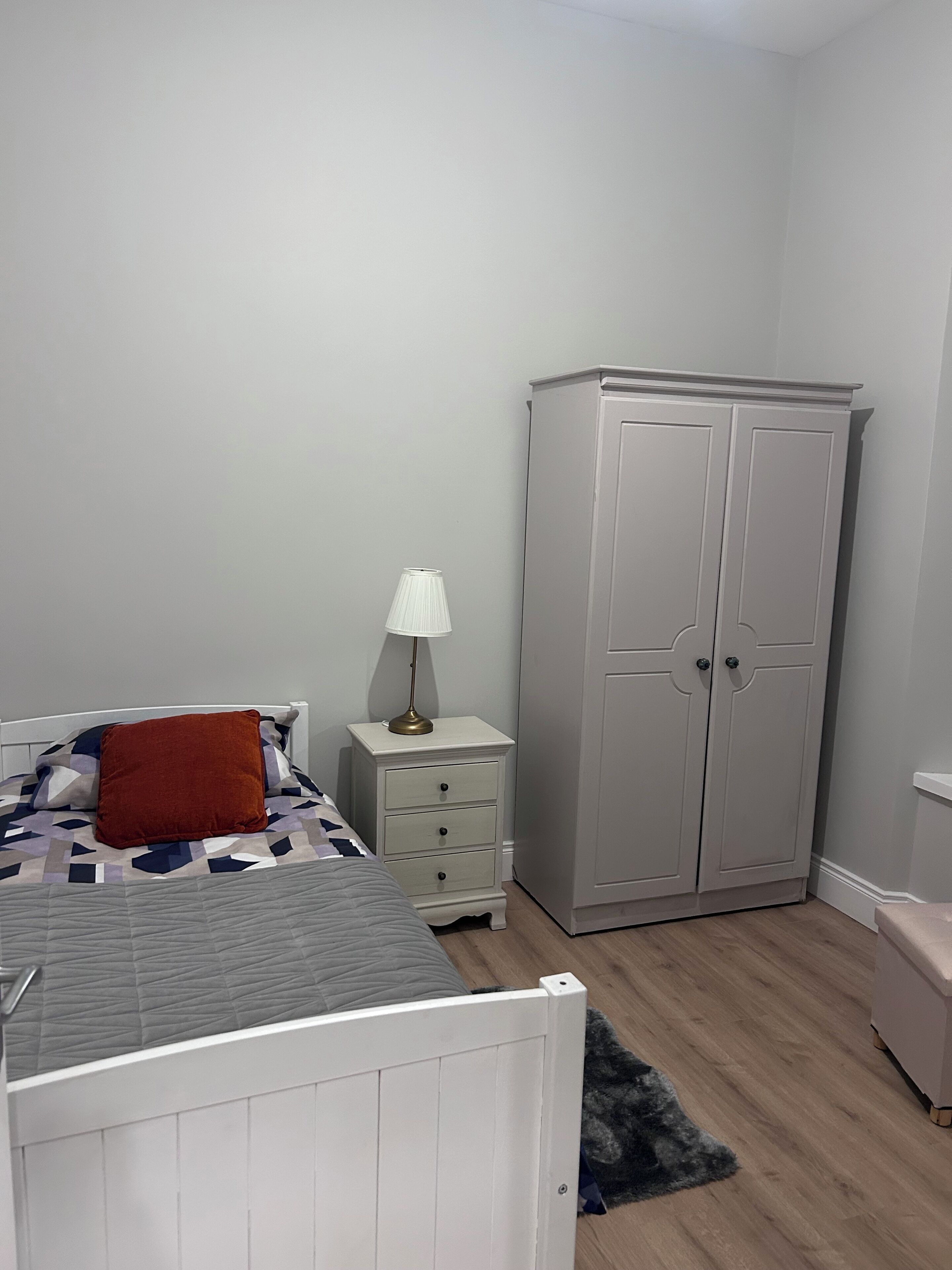 A bedroom with a bed, nightstand, lamp, wardrobe, and a small bench.