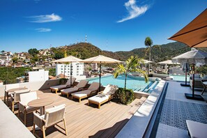 A rooftop pool area with lounge chairs, umbrellas, and a view of a hillside town.