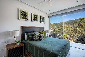 A bedroom with a large bed, a nightstand, a lamp, and a view of a mountainous landscape through a balcony.