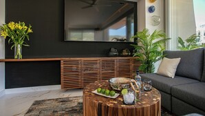 Una sala moderna con un televisor de pantalla plana grande montado en una pared oscura, una mesa consola de madera con objetos decorativos, una mesa redonda de centro con bebidas y lima, y un sofá gris.