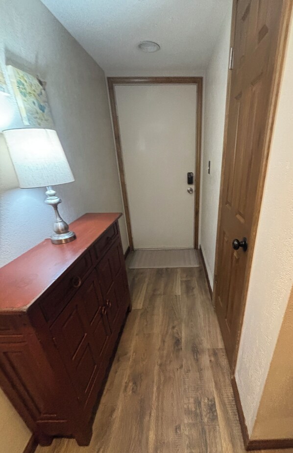Un couloir étroit avec un plancher en bois, une porte blanche et une commode en bois sur laquelle est posée une lampe.