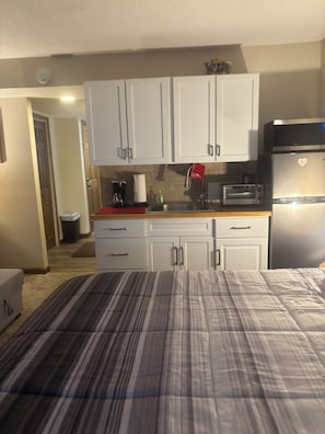 A compact hotel room with a kitchenette, a bed with striped bedding, and a view of the hallway.