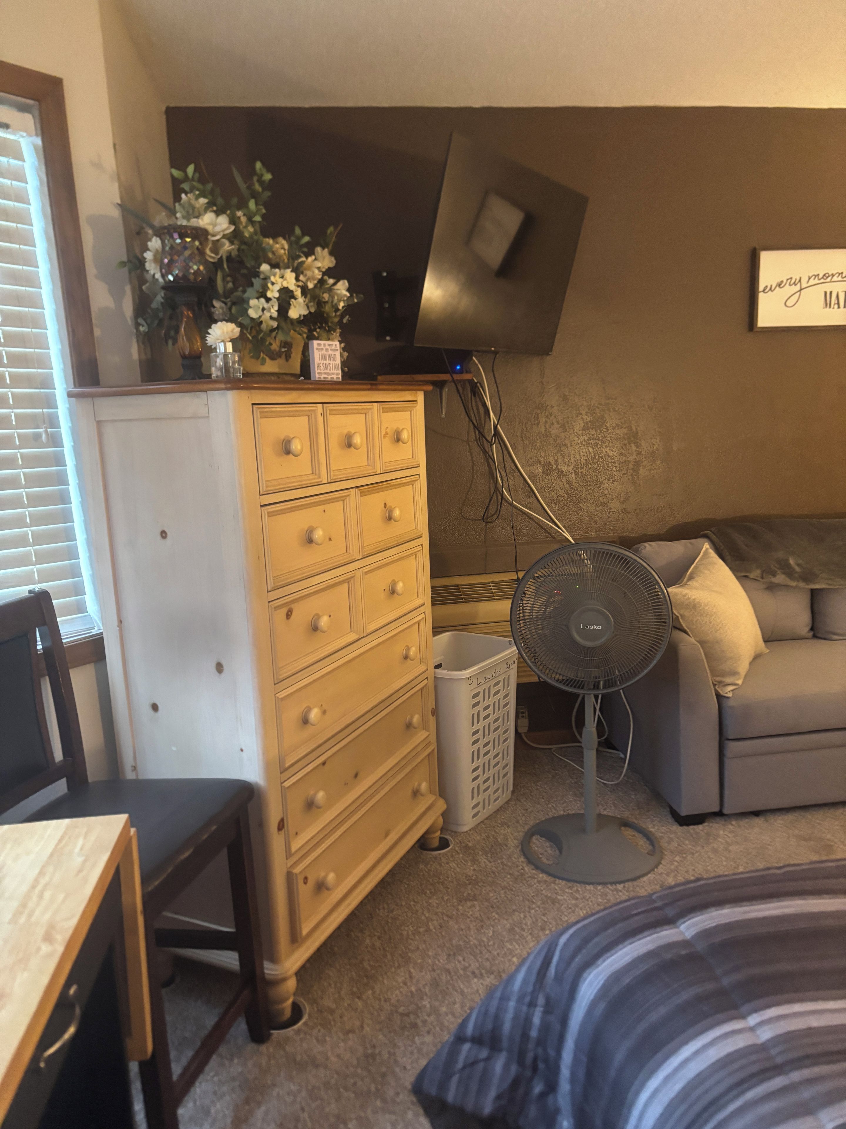 Quarto com cômoda de madeira, cesto de roupa branca, sofá cinza e ventilador.
