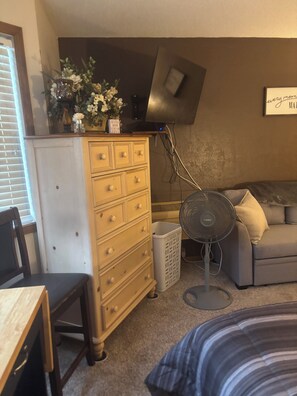 Une pièce comprenant une commode en bois, un panier à linge blanc, un canapé gris et un ventilateur sur pied.