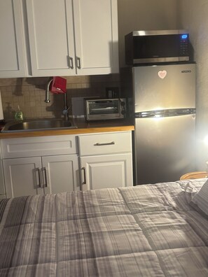 A kitchenette with a toaster, a microwave, and a refrigerator. There is a sink with a faucet and a red towel hanging on it. The room has white cabinets and a bed with a striped blanket.