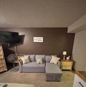 Un soggiorno con un divano modulare grigio, una TV a schermo piatto montata a parete, una lampada da terra e un comodino con una lampada e fazzoletti.