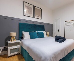 A neatly made bed with a teal headboard, white bedding, and two pillows. There are two bedside tables with lamps, a framed artwork above the bed, and a door to the right. - Modern Townhouse-free Parking-game Room-w-ifi (Manchester)