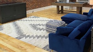 A living room with a brick wall, wooden beams, a flat-screen TV, a pool table, blue sofas, and a patterned rug. - Market street apartment (Corning)