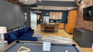 A pool table in a modern living room with a blue sofa, a kitchen in the background, and a brick wall. - Market street apartment (Corning)