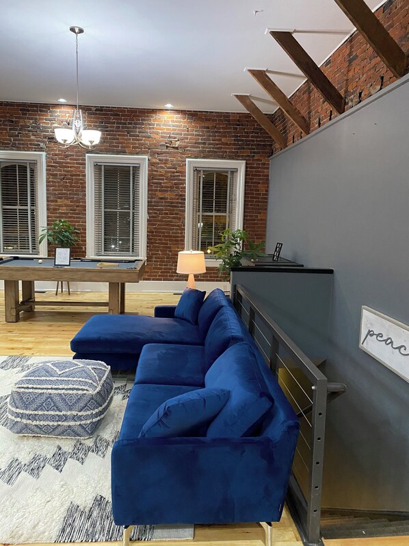 A modern living room with a blue sectional sofa, a dining table with chairs, and a brick wall.