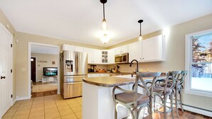 A modern kitchen with white cabinets, a central island with bar stools, stainless steel appliances, and a large window providing natural light.