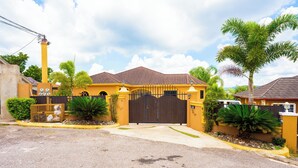 A yellow house with a brown gate, surrounded by greenery and a paved driveway. - Cozy 1-bedroom apartment in charming Mandeville with WiFi, AC (Mandeville)