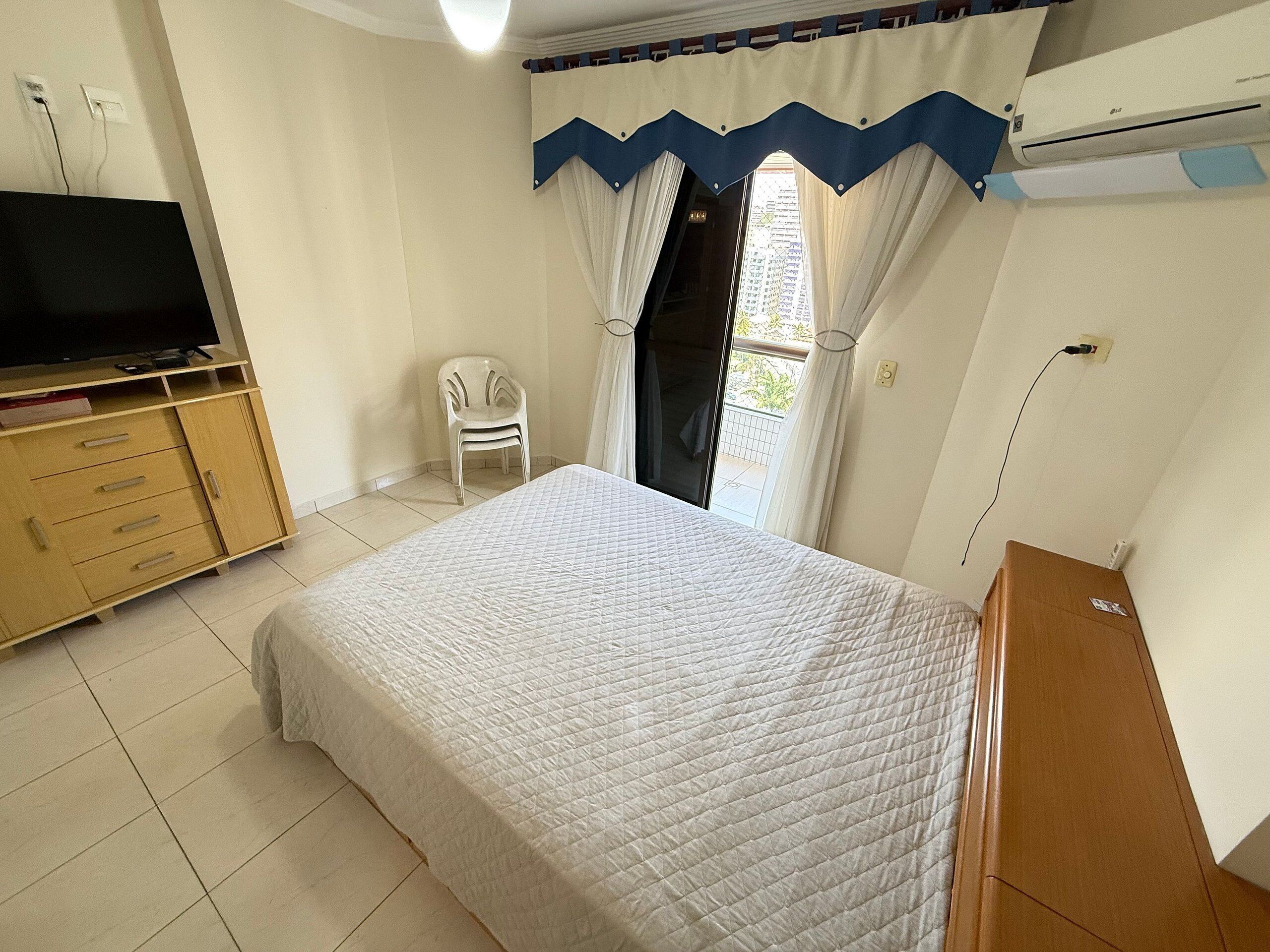 Un dormitorio con cama, televisor sobre un mueble de madera, una silla y una ventana con cortinas.