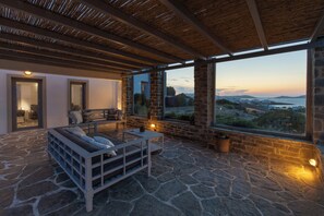 Patio con suelo de piedra, sofá y mesa, ventanas amplias con vistas al paisaje y estructura de techo de madera.