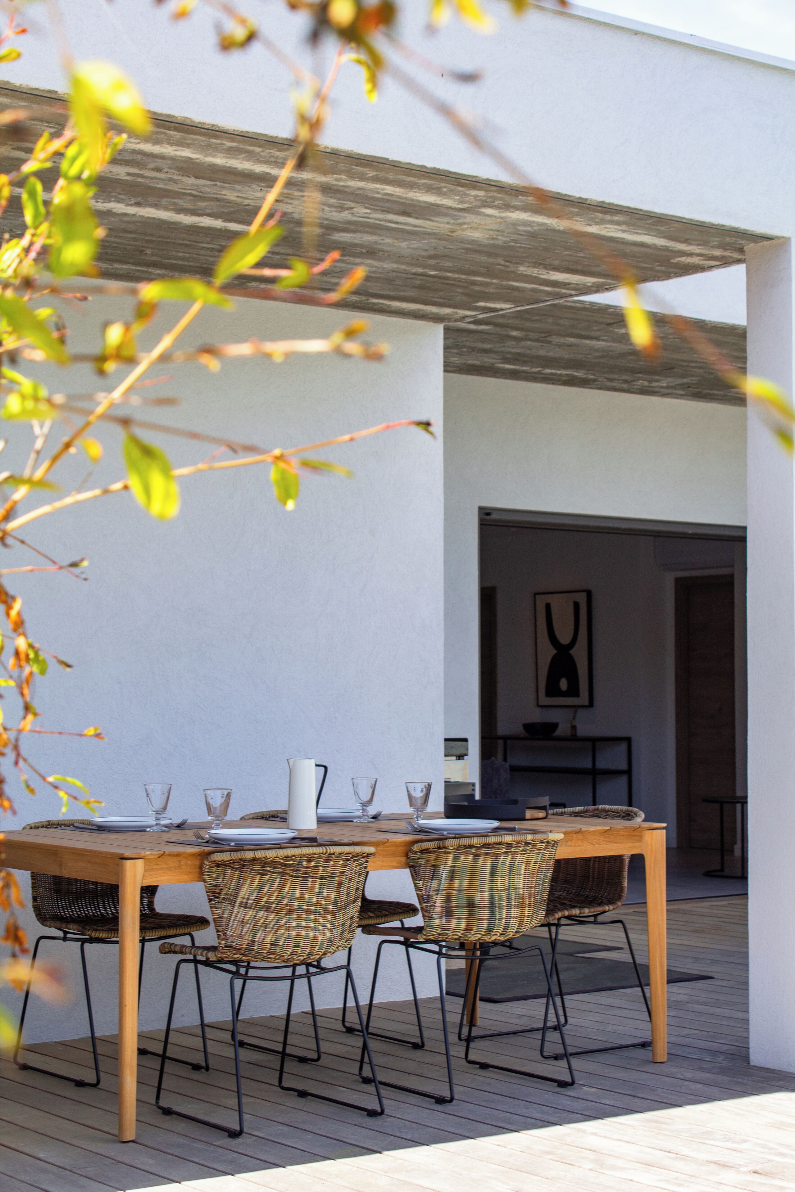 An outdoor dining area with a wooden table and wicker chairs, a white pitcher, and wine glasses.