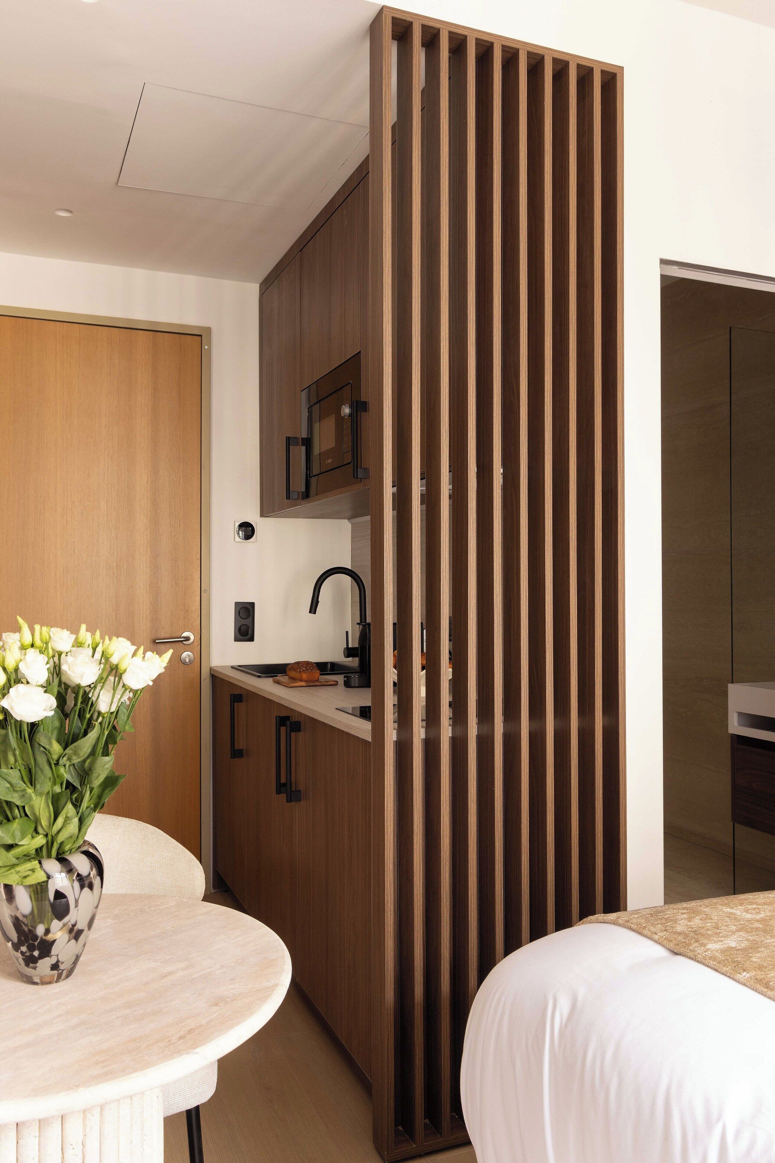 Una camera d'albergo moderna con un divisorio in legno, un angolo cottura, un tavolo rotondo con un vaso di fiori e un letto con copriletto color beige.
