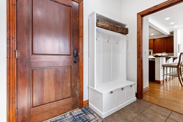 Una puerta de madera con un armario blanco y un banco, una alfombra, y una cocina visible al fondo.