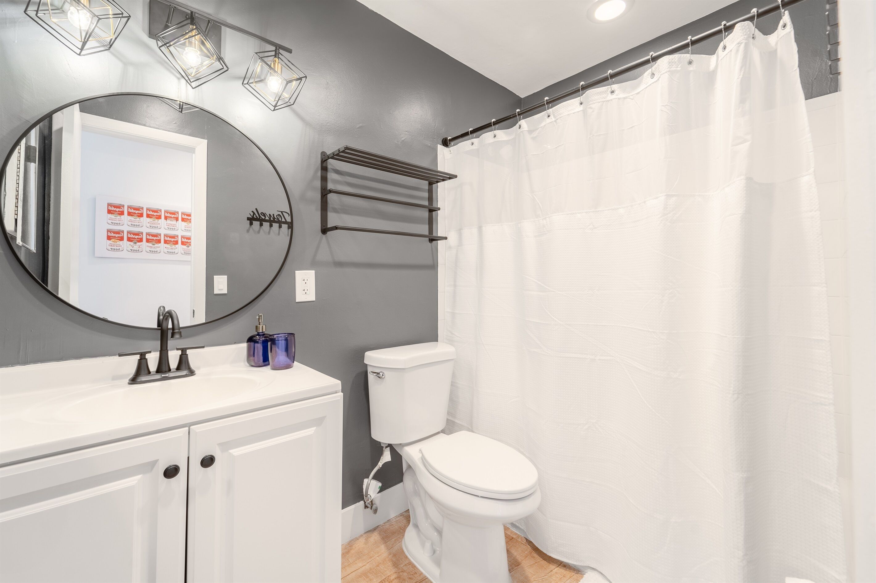 Une salle de bain avec des toilettes blanches, un miroir rond, un lavabo blanc et un rideau de douche.